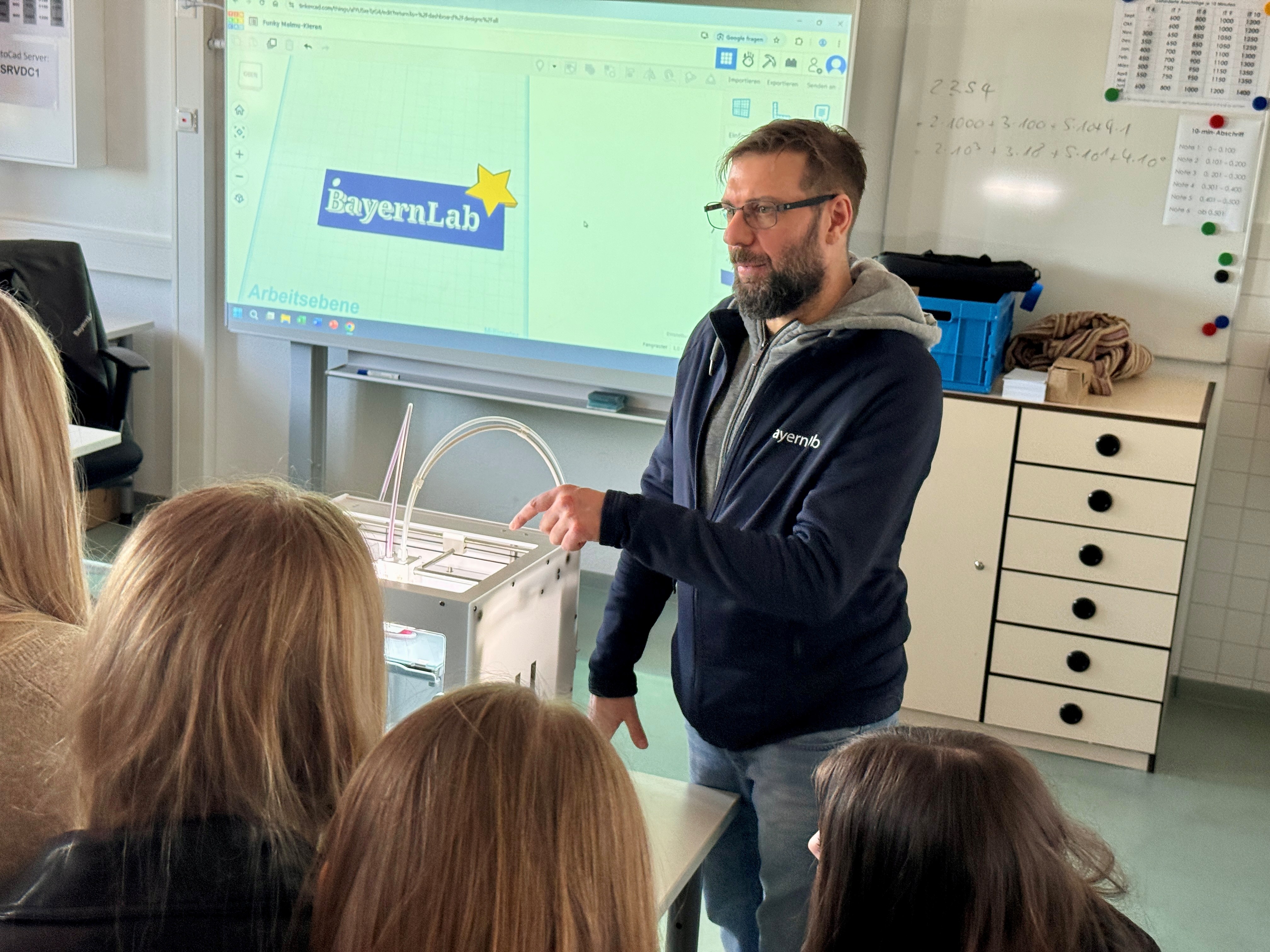 Ein Mann mit Bart steht in einem Klassenraum und zeigt auf einen 3D-Drucker. Vor ihm stehen Mädchen, von denen man nur die Hinterköpfe sieht. 